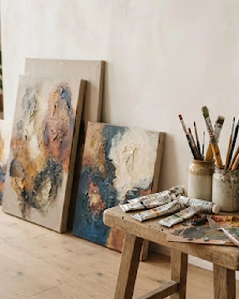 Sunlit studio corner with textured abstract paintings leaning against a wall beside brushes and paint
